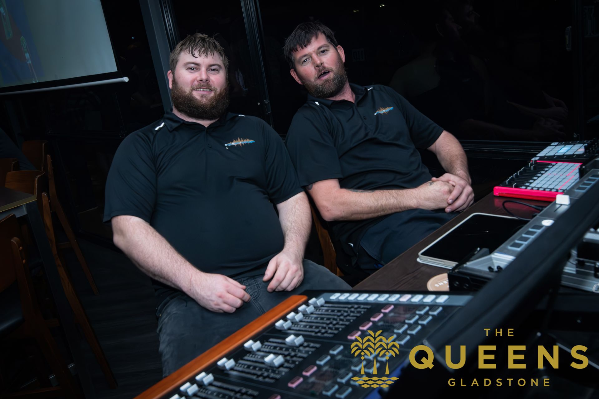 Two men sitting behind a DJ booth in a dark venue, with equipment and “The Queens Sladstone” text visible