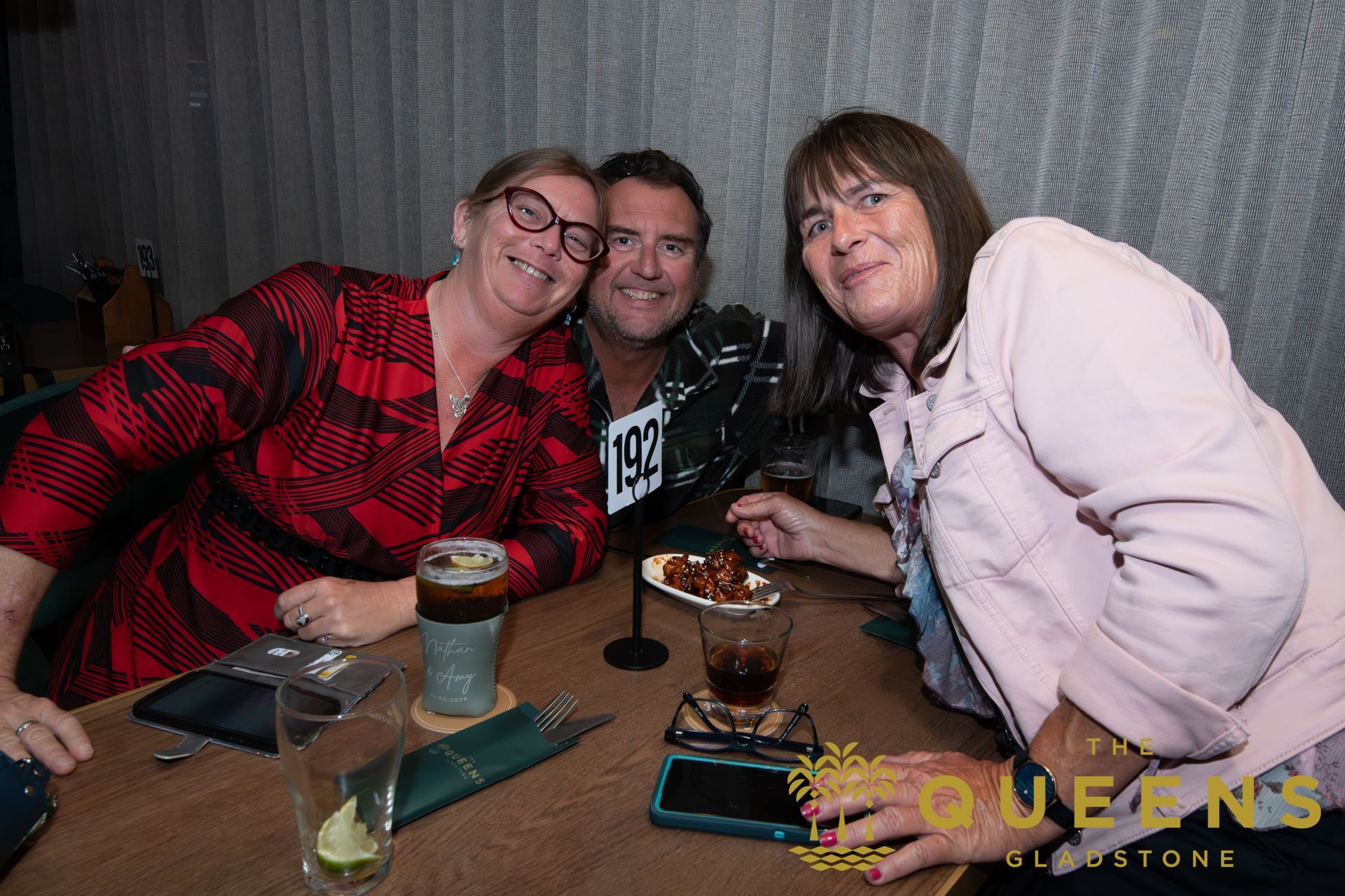 Three people smiling at a table with drinks in a dimly lit restaurant, posing close together.