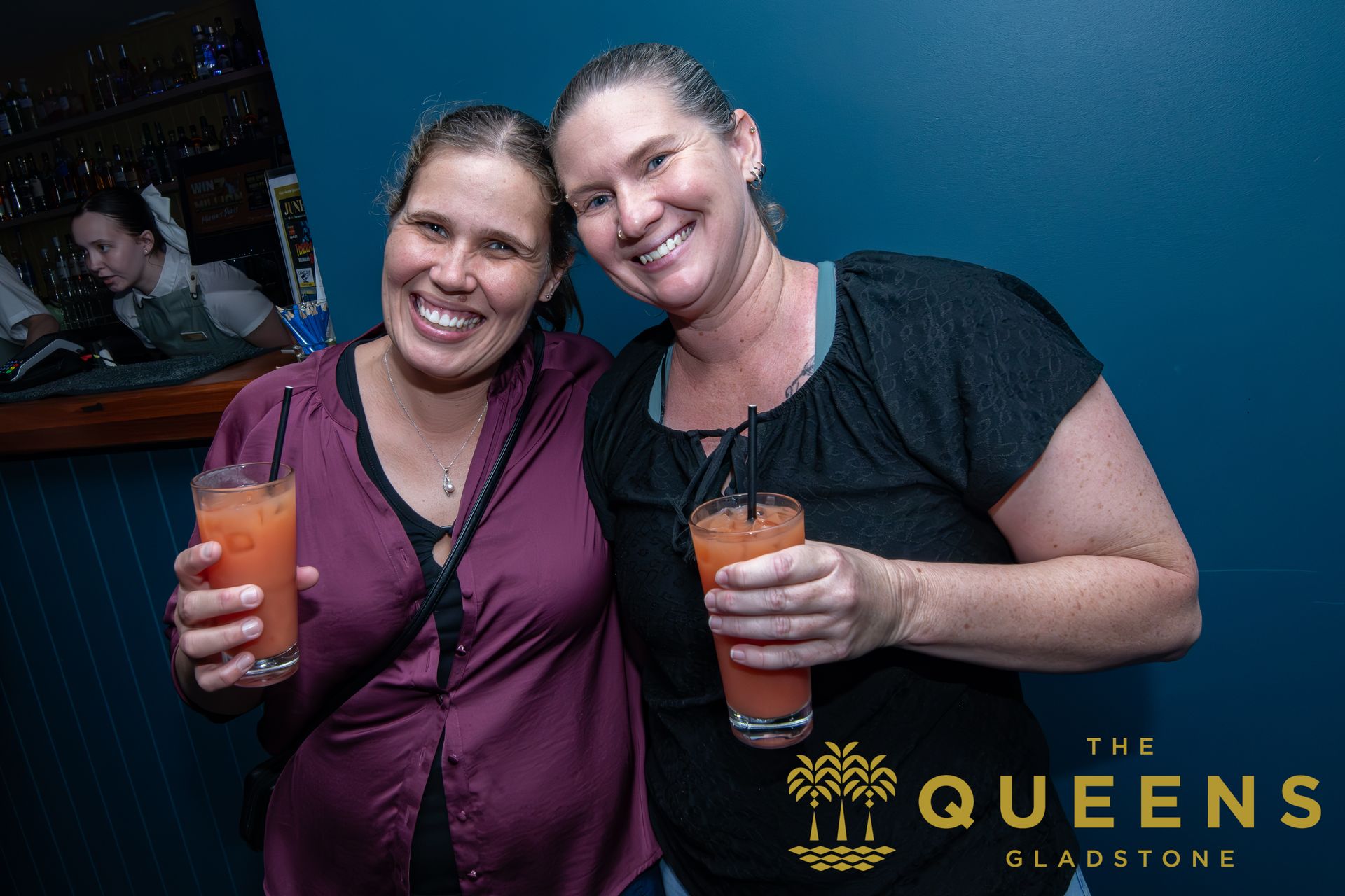 Two women smiling and holding cocktails in a nightclub, with The Queens Gladstone logo in the corner