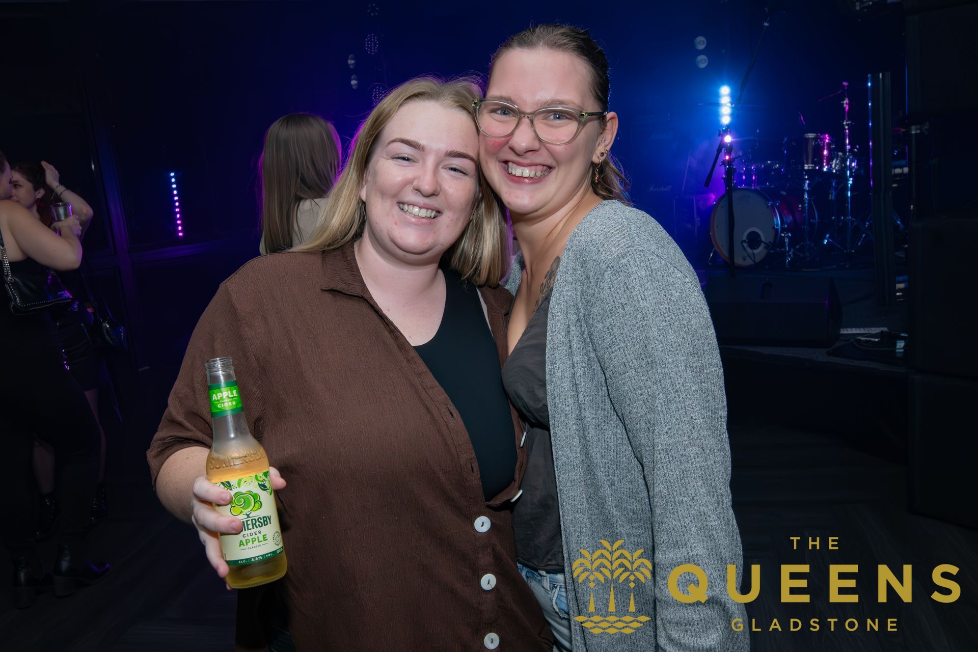 Two smiling women pose at a nightclub, one holding a beer bottle under purple stage lights.