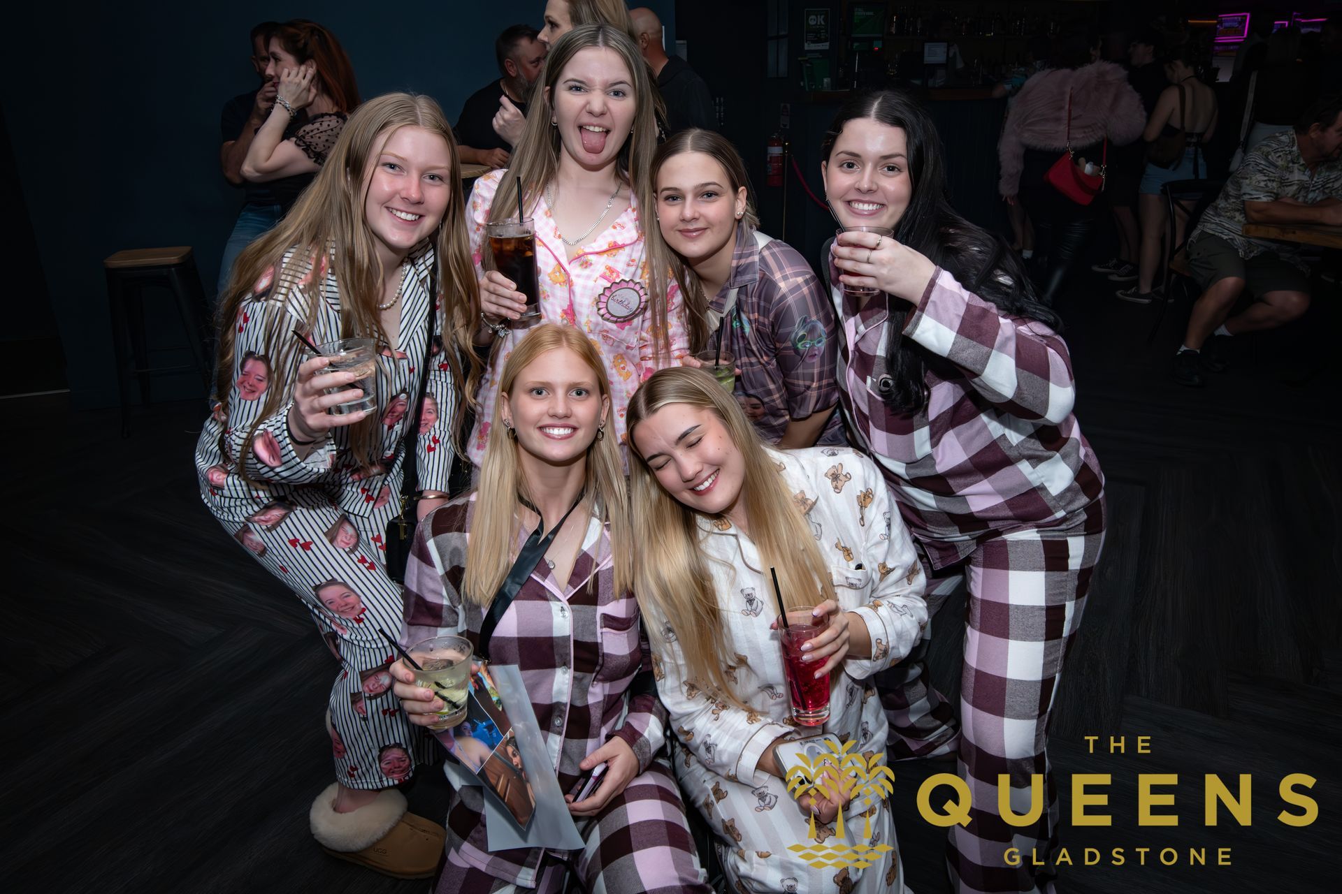 Six women pose smiling in matching plaid outfits at an event, with a “The Queens” logo in the corner