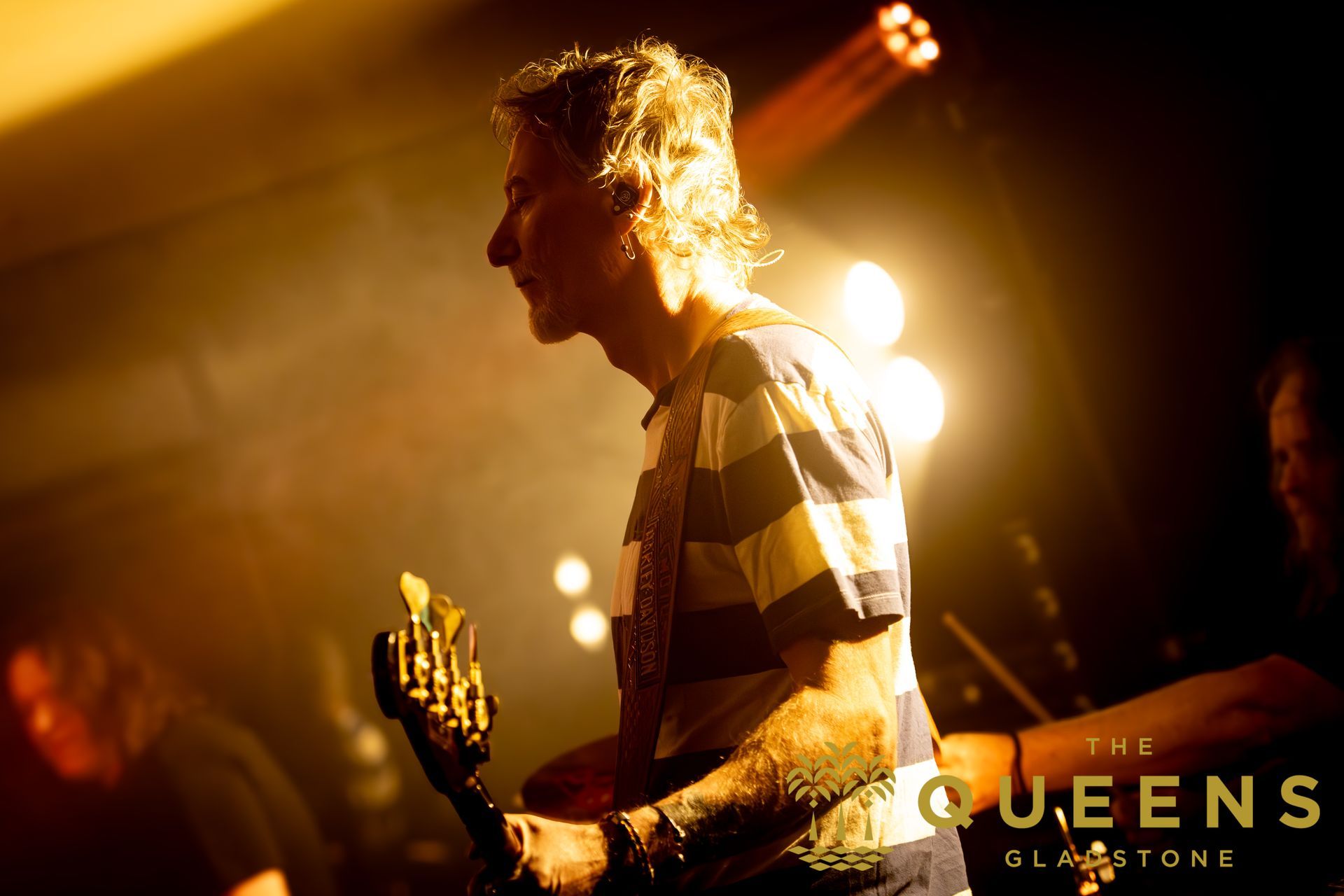 Silhouetted guitarist onstage under warm orange lights, with the band name “QUEENS” visible in the corner