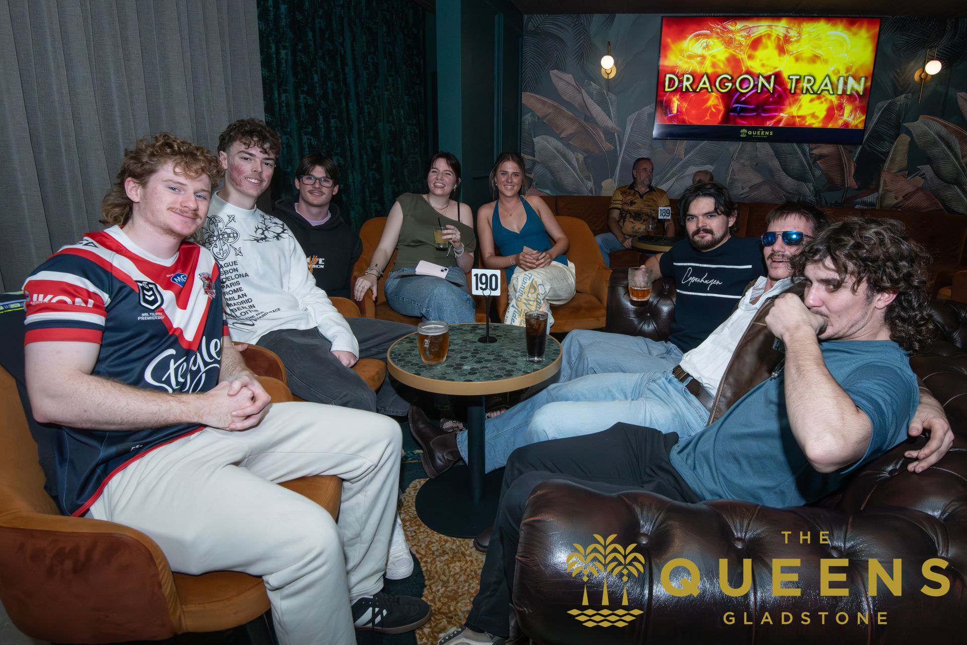 People relaxing on lounge chairs in a dim gaming lounge with a neon screen and a “The Queens” sign