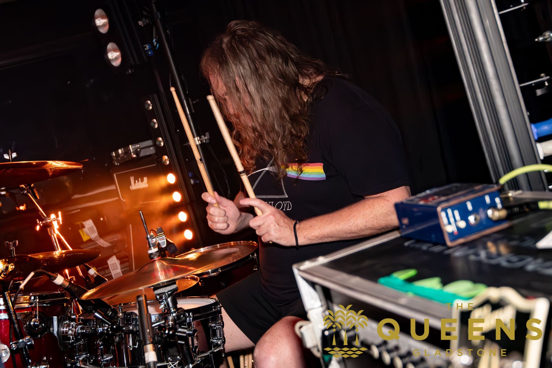 Drummer playing on stage under warm lights, wearing a black shirt with a rainbow stripe.
