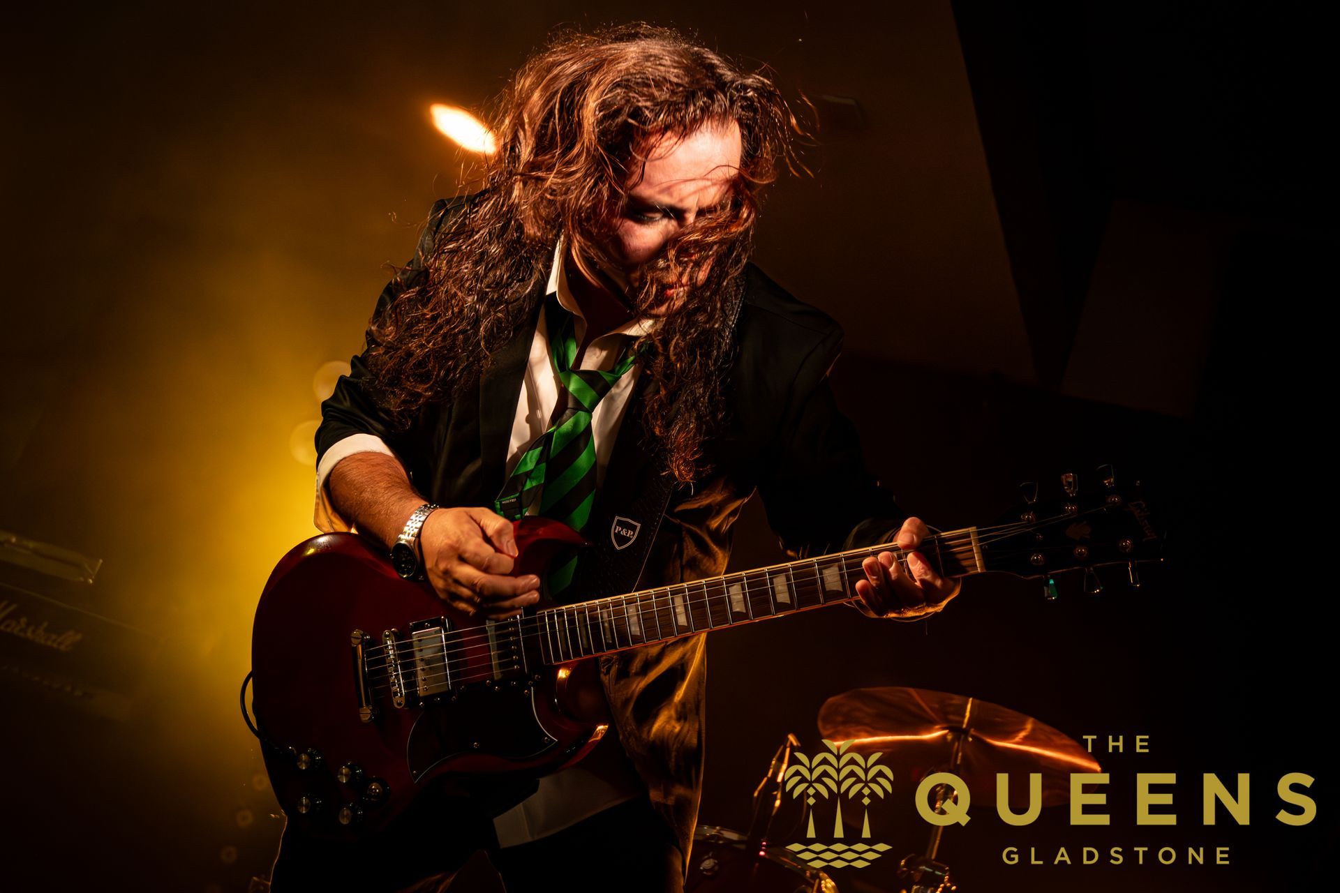 Guitarist performing under warm stage lights, with “The Queens of Sladstone” text visible in the corner.