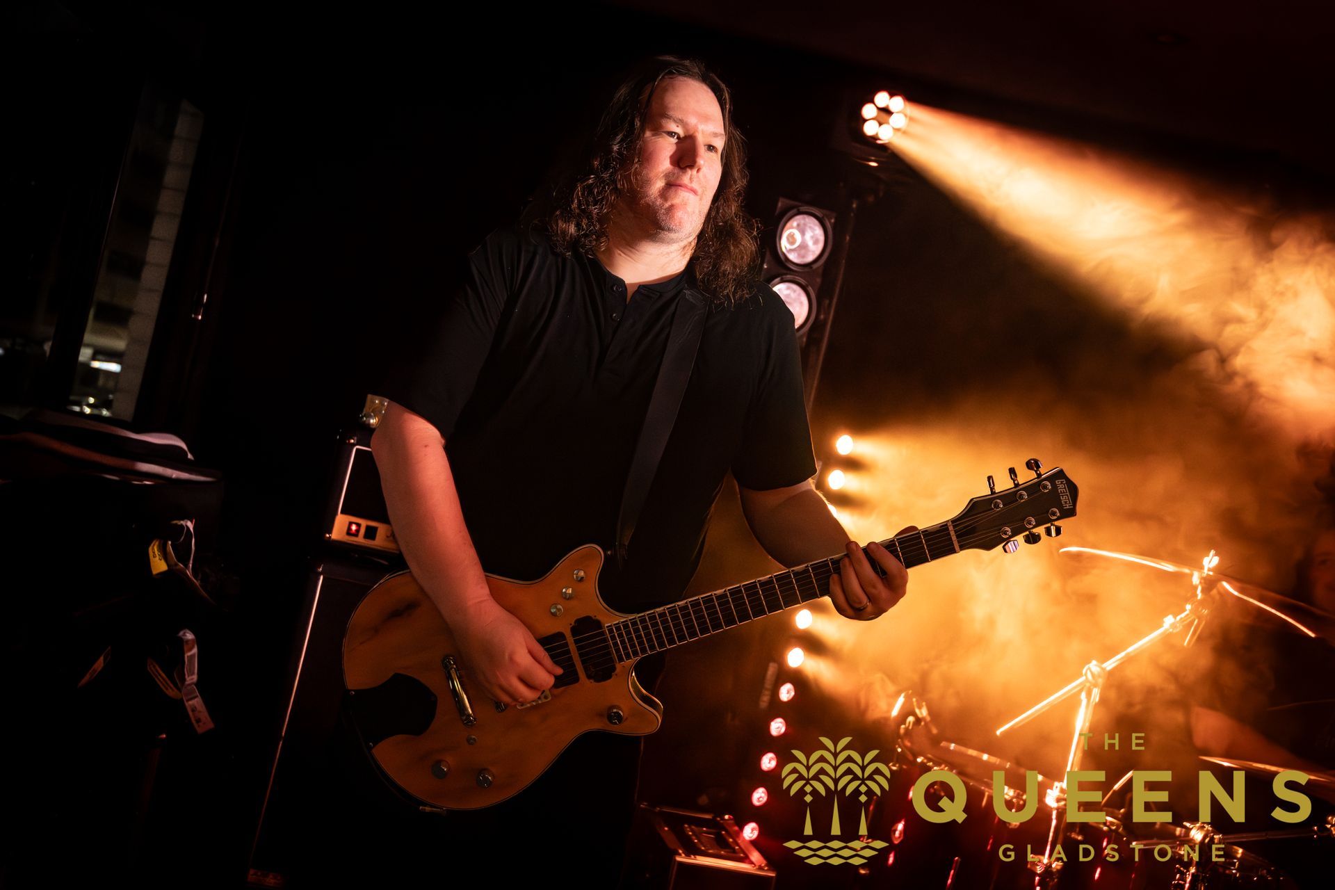 Guitarist onstage under warm orange lights, playing an electric guitar with “QUEENS” visible in the background.