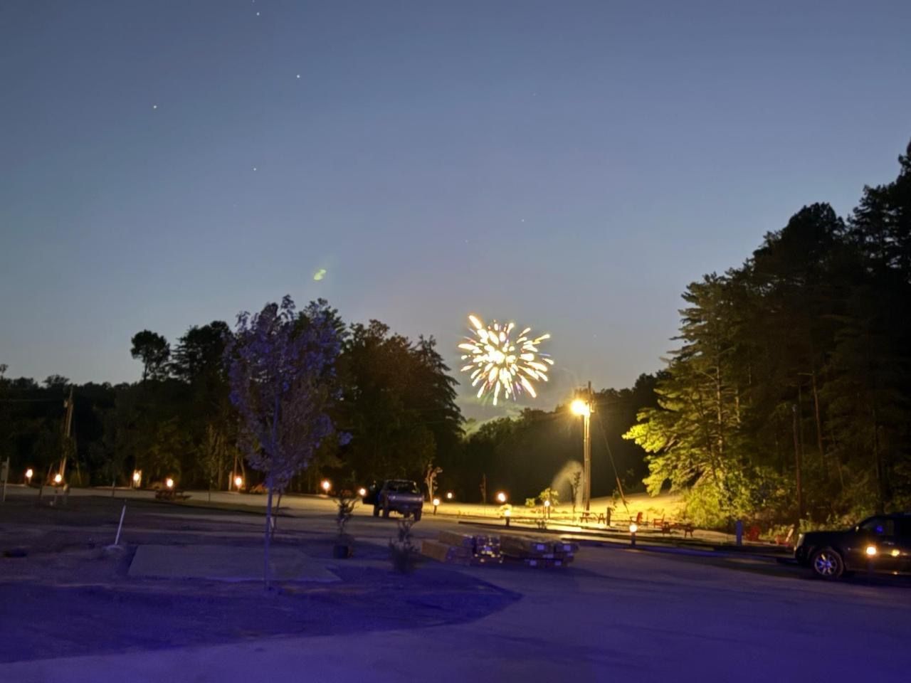 Fireworks explode over a dark treeline and parking lot at dusk.