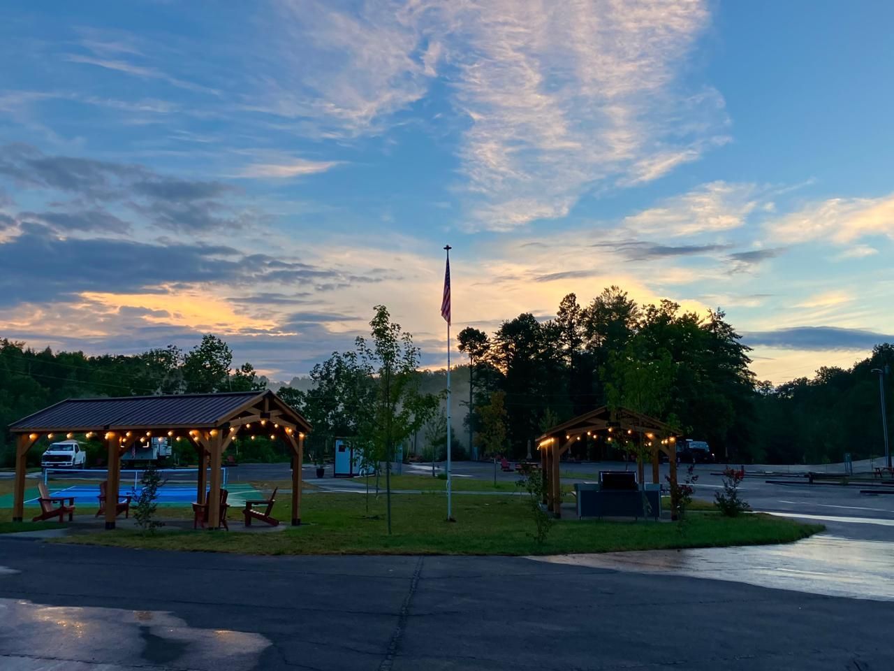 Evening view of a park with string lights, gazeboes, American flag, and cloudy sky.