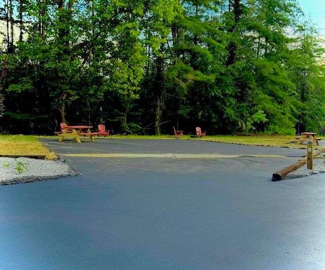 Asphalt parking area with picnic tables and Adirondack chairs, surrounded by trees.