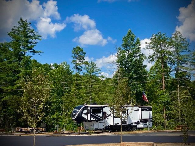 RV parked at a campsite, American flag waving. Lush green trees and a partly cloudy blue sky.