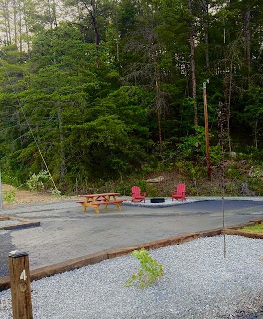 Campsite with picnic table, red chairs, fire pit, and gravel area, backed by a forest.
