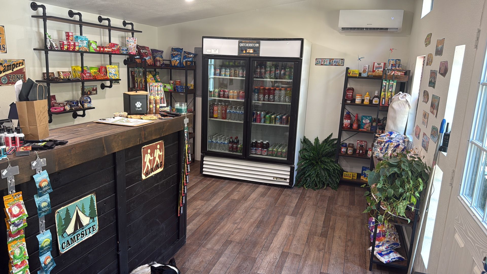 Small convenience store interior with snacks, drinks, cooler, and a counter with a register.