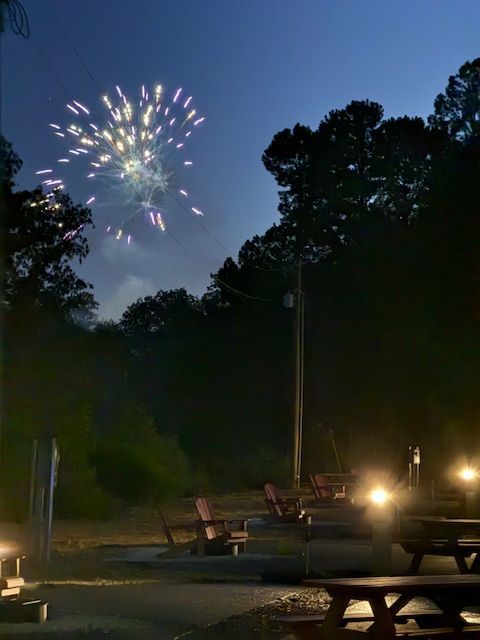 Fireworks burst over a dark campground with picnic tables and Adirondack chairs.