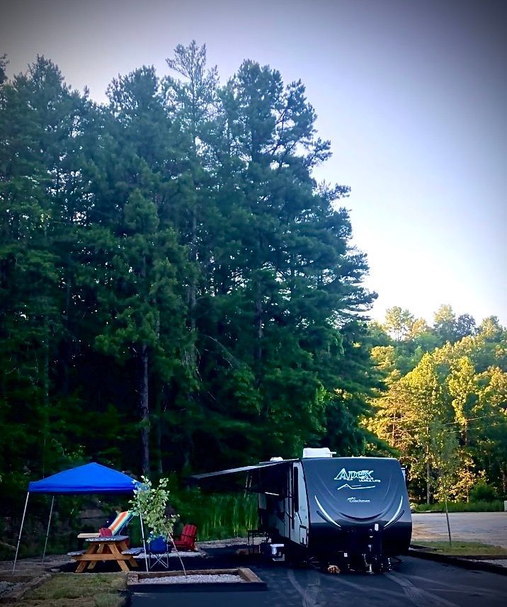 RV parked at a campsite with a blue canopy and a forested background on a sunny day.