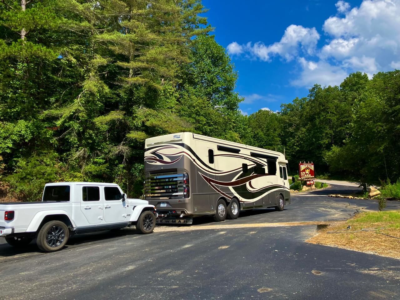 White truck towing a large RV on a paved road, surrounded by trees under a blue sky.