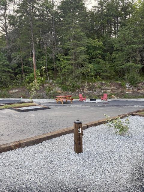 Campground site with gravel, wooden picnic table, red chairs, and trees.