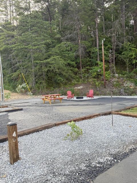 Campsite with picnic table, fire pit, red chairs, gravel, and trees in the background.