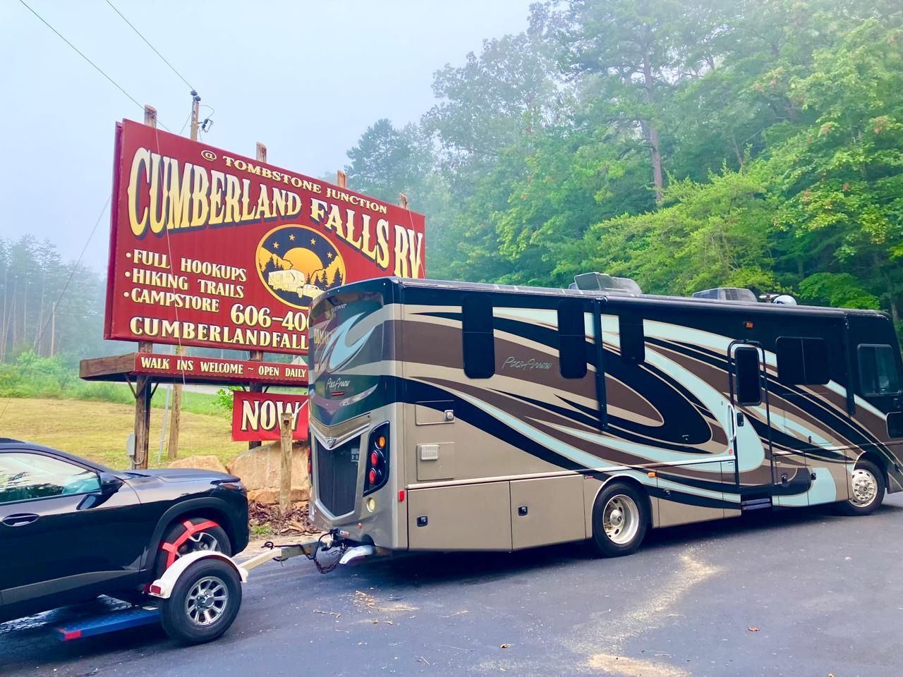 RV parked at Cumberland Falls RV park entrance sign. Grey RV with a black car trailer.