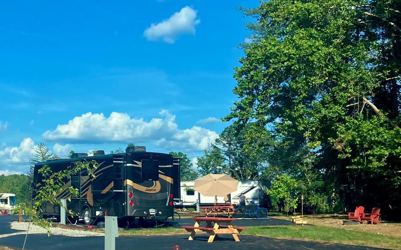 RV parked at a campsite with picnic table, umbrella, and trees under a blue sky.