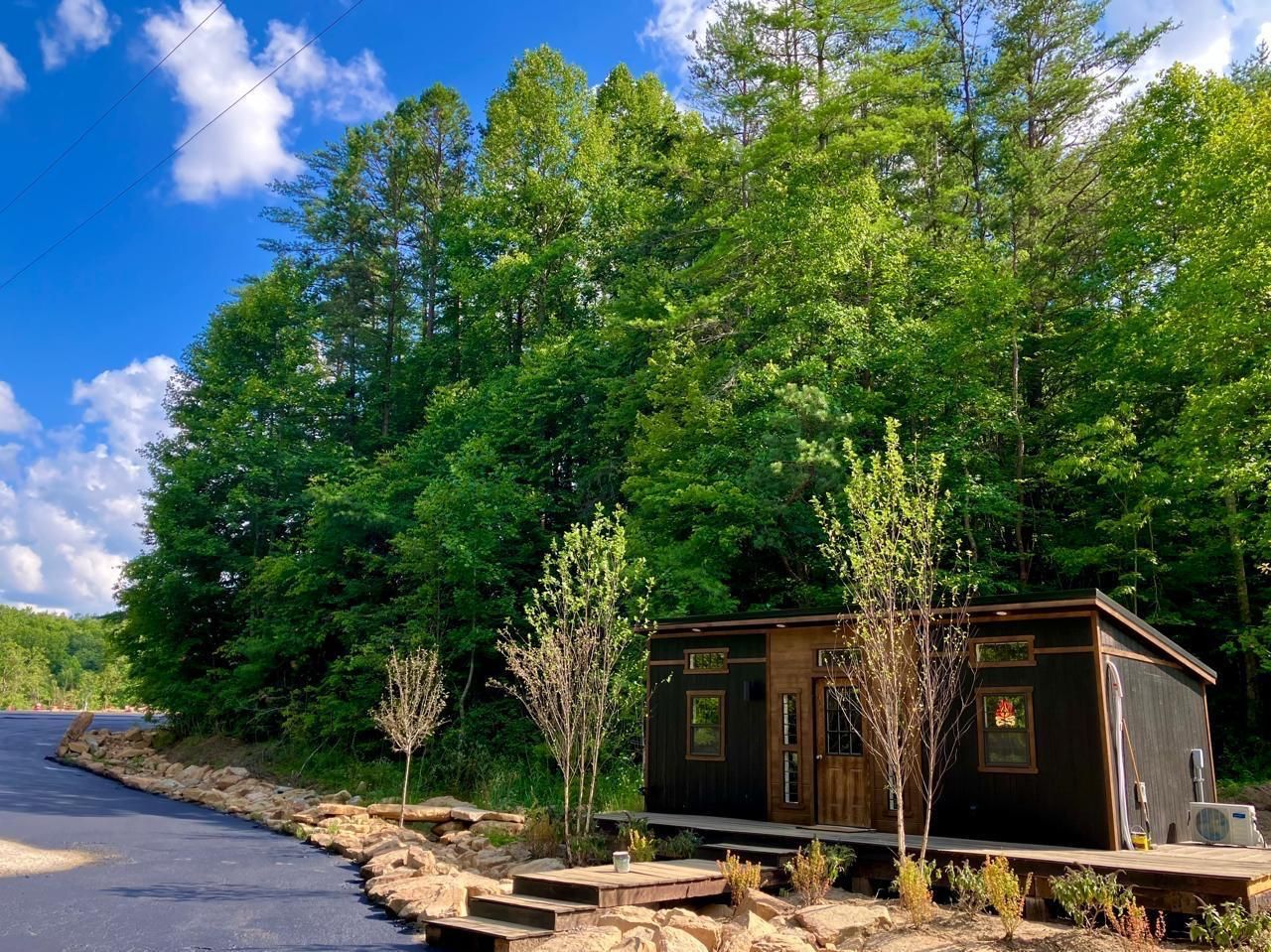 A small dark cabin sits nestled among lush green trees under a blue sky.
