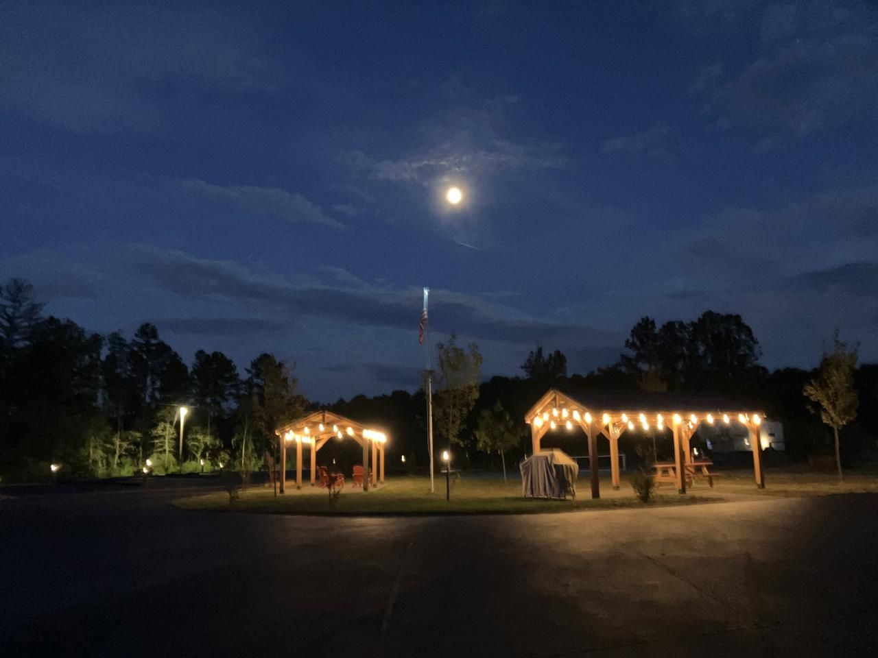 Night scene: illuminated gazebos, string lights, and full moon in a dark blue sky.