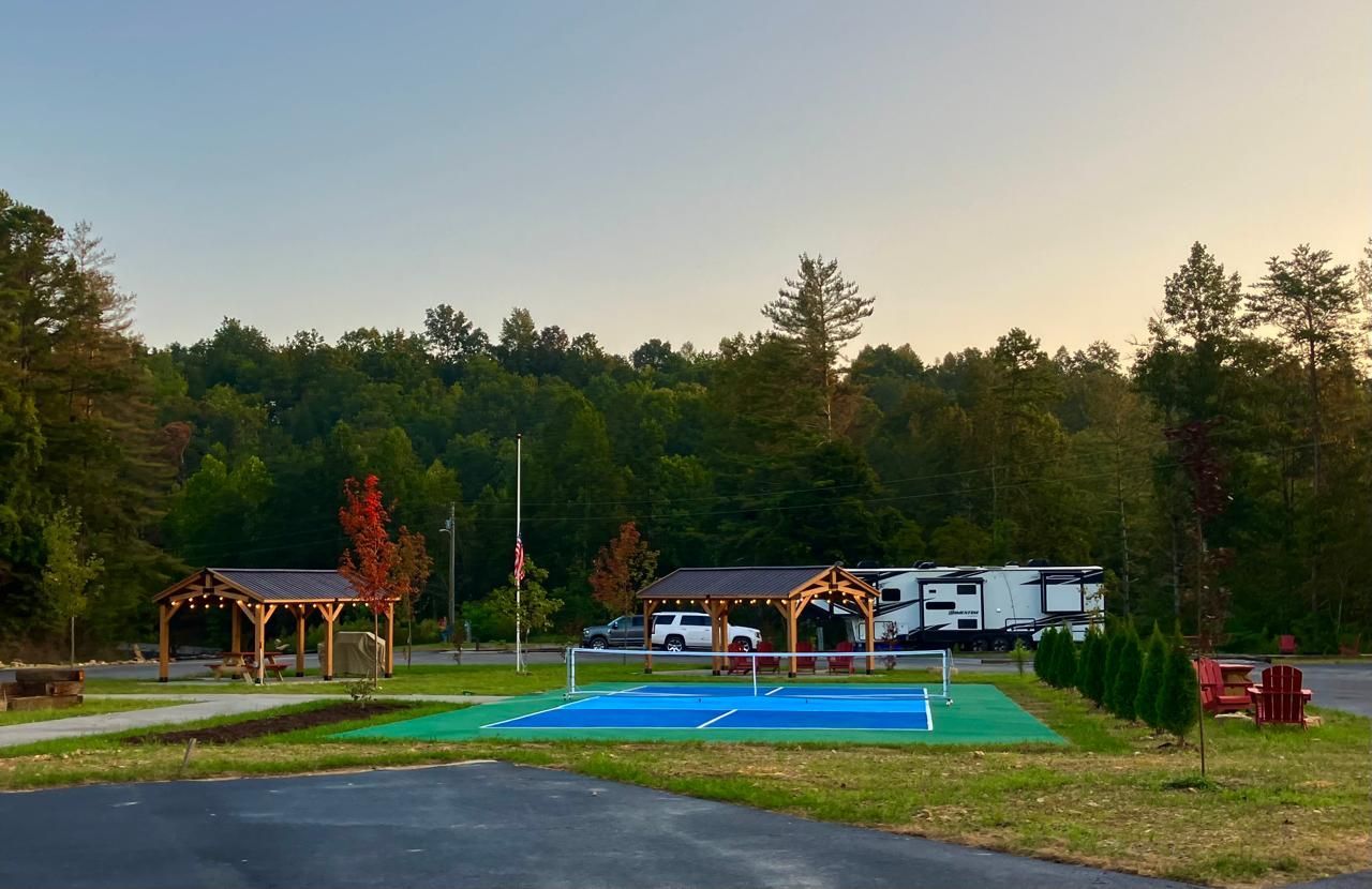 Campground scene: RV parked near a covered picnic area, court, trees. Early morning light.