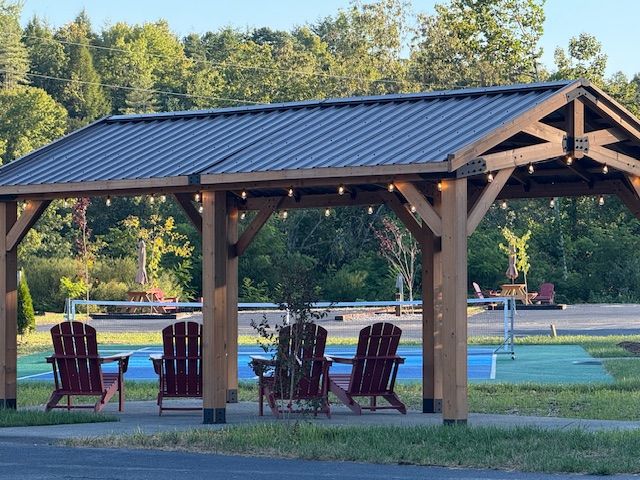 Wooden gazebo with string lights, red Adirondack chairs, and blue/green pavement.