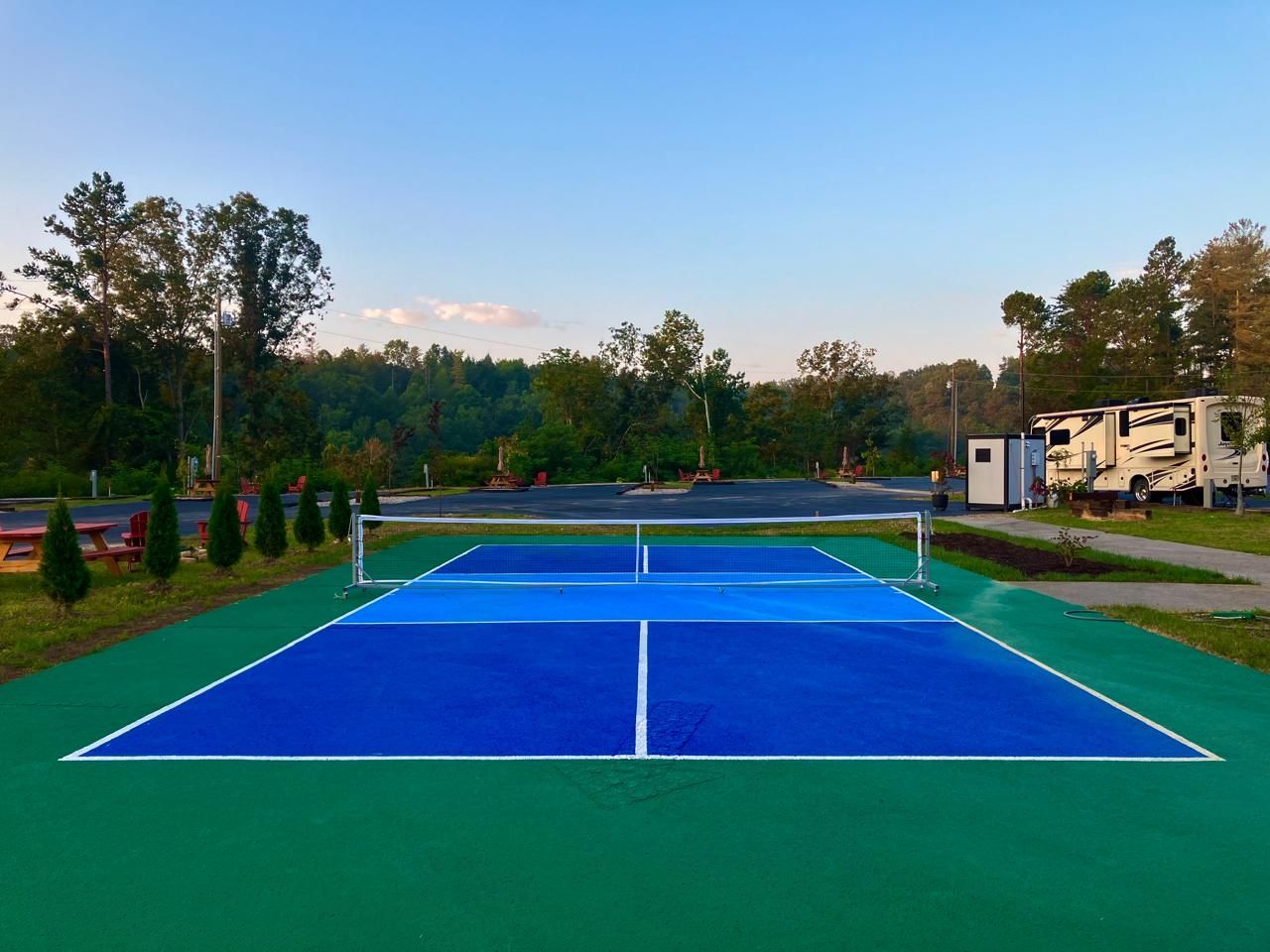 A pickleball court with blue playing surface, white net, and green surroundings, set outdoors with RVs and trees.