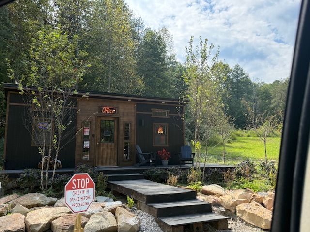 Tiny dark cabin with wooden door, rock steps, surrounded by trees and a lawn. 