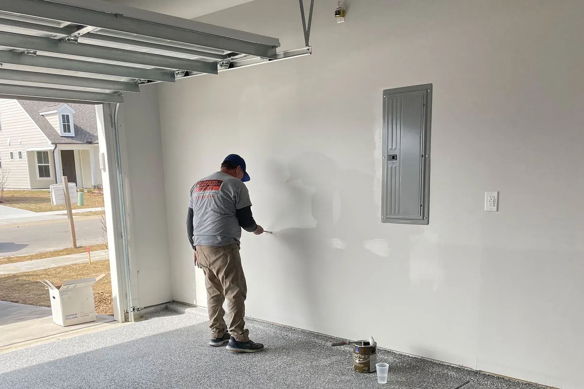 Man painting a white wall in a garage, next to a metal electrical panel. Gray floor, open doorway.