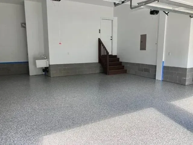 Empty garage with a speckled gray floor and white walls. A door and stairs are visible in the background.