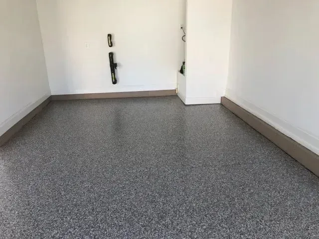 Empty garage with speckled gray floor, white walls, and brown baseboards.