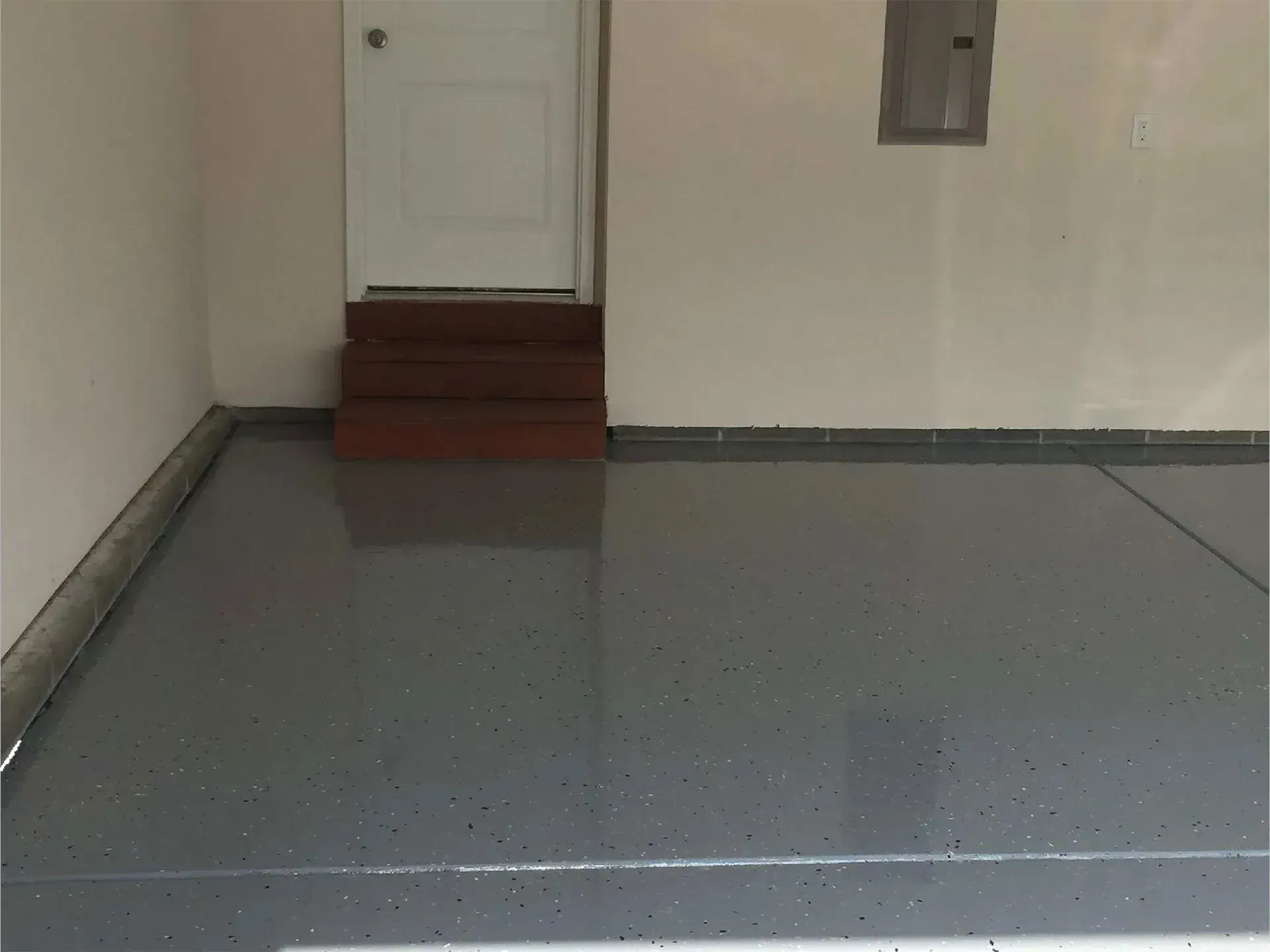 Grey epoxy-coated garage floor with steps leading to a white door. Concrete walls and a grey baseboard.