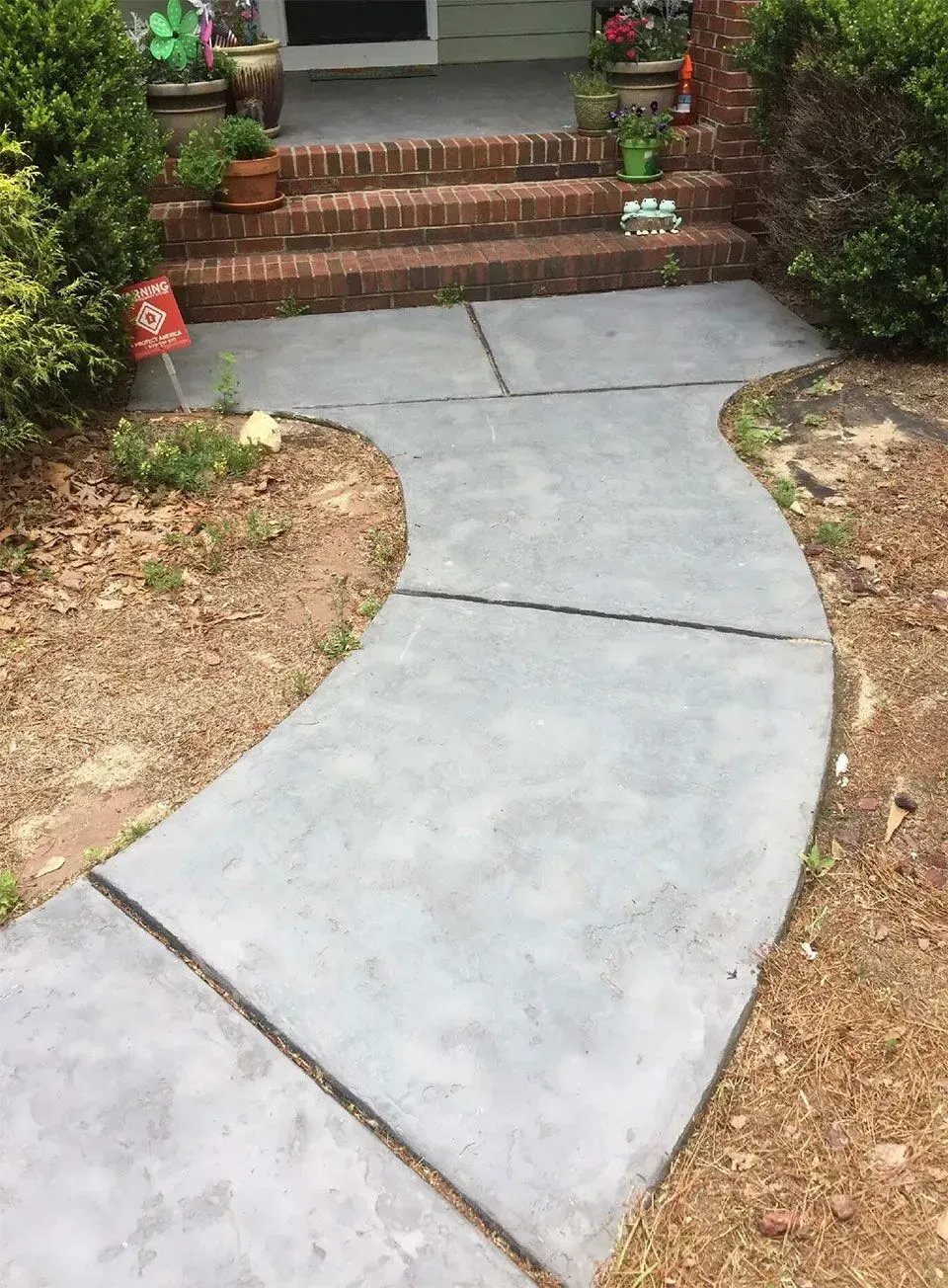 Gray concrete walkway curves to a brick-stepped entrance of a house, surrounded by dirt and greenery.