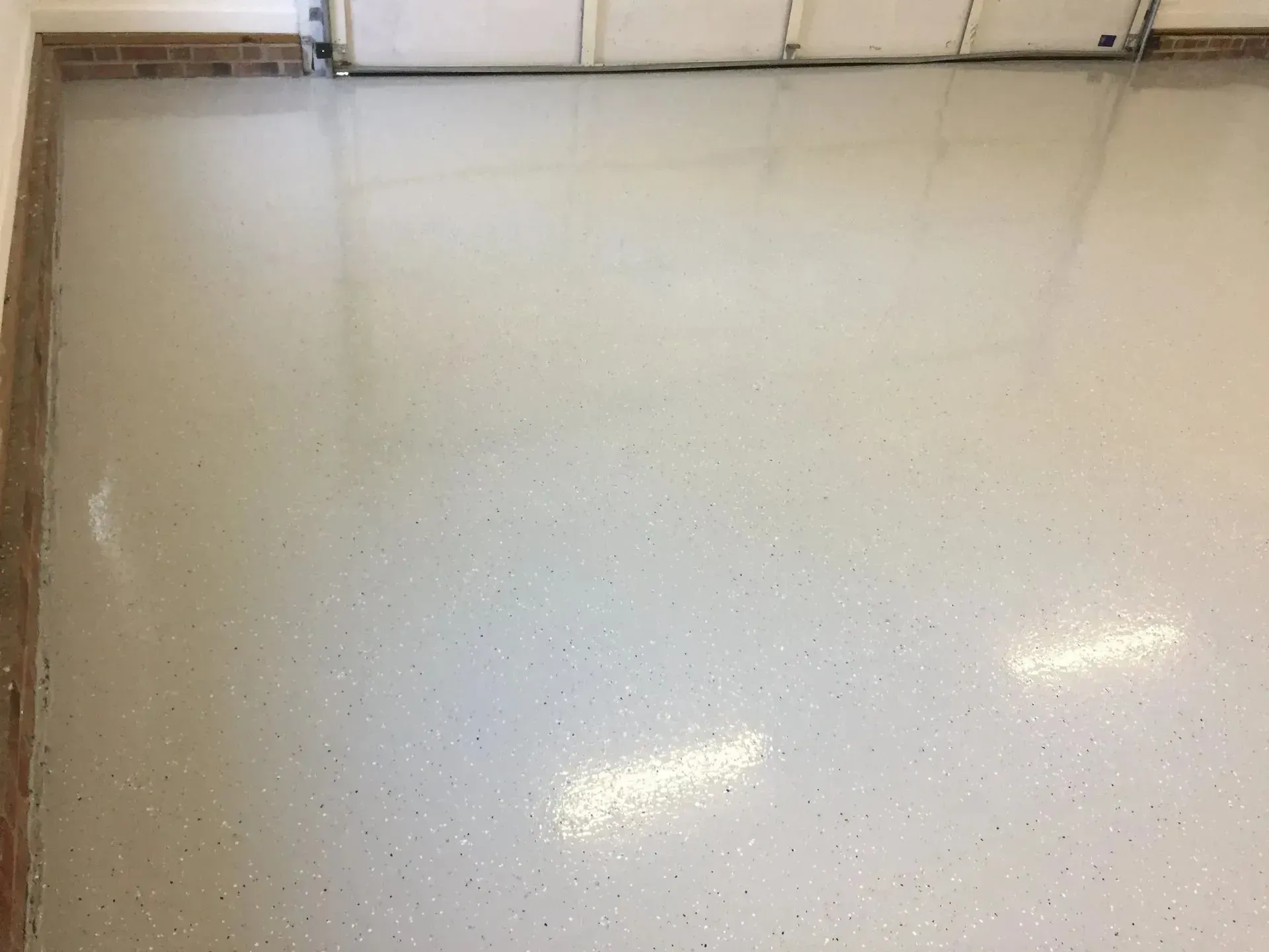 Shiny, speckled, light-colored garage floor reflecting light. Brick wall on the left, garage door at top.