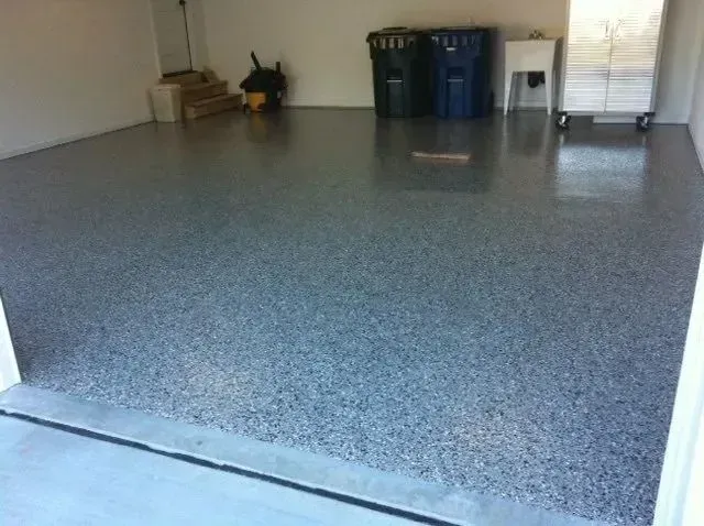 Garage with speckled gray epoxy floor. Black trash cans and a white cabinet against the wall.