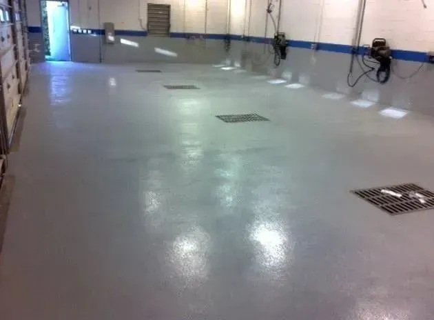 Empty, gray-floored automotive shop with drains, overhead equipment, and a blue trim. Open door to the left.