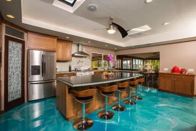 Kitchen with blue epoxy floor, dark island with seating, stainless steel appliances, and wood cabinets.
