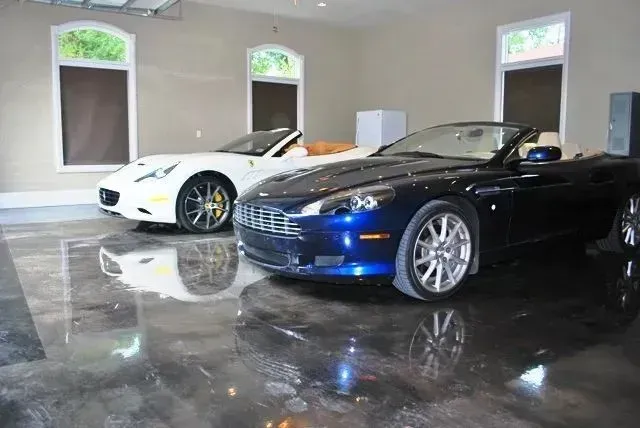 Two luxury cars, a white Ferrari and a blue Aston Martin, parked in a polished garage with arched windows.