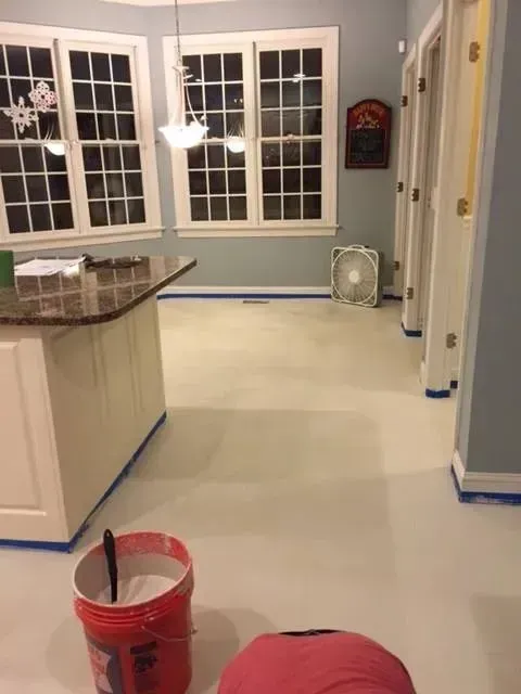 Kitchen with freshly painted light floor, white cabinets, and blue walls. Red bucket, tape, and fan visible.