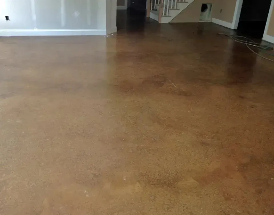 Brown stained concrete floor in a room, reflecting light. White walls and staircase in background.