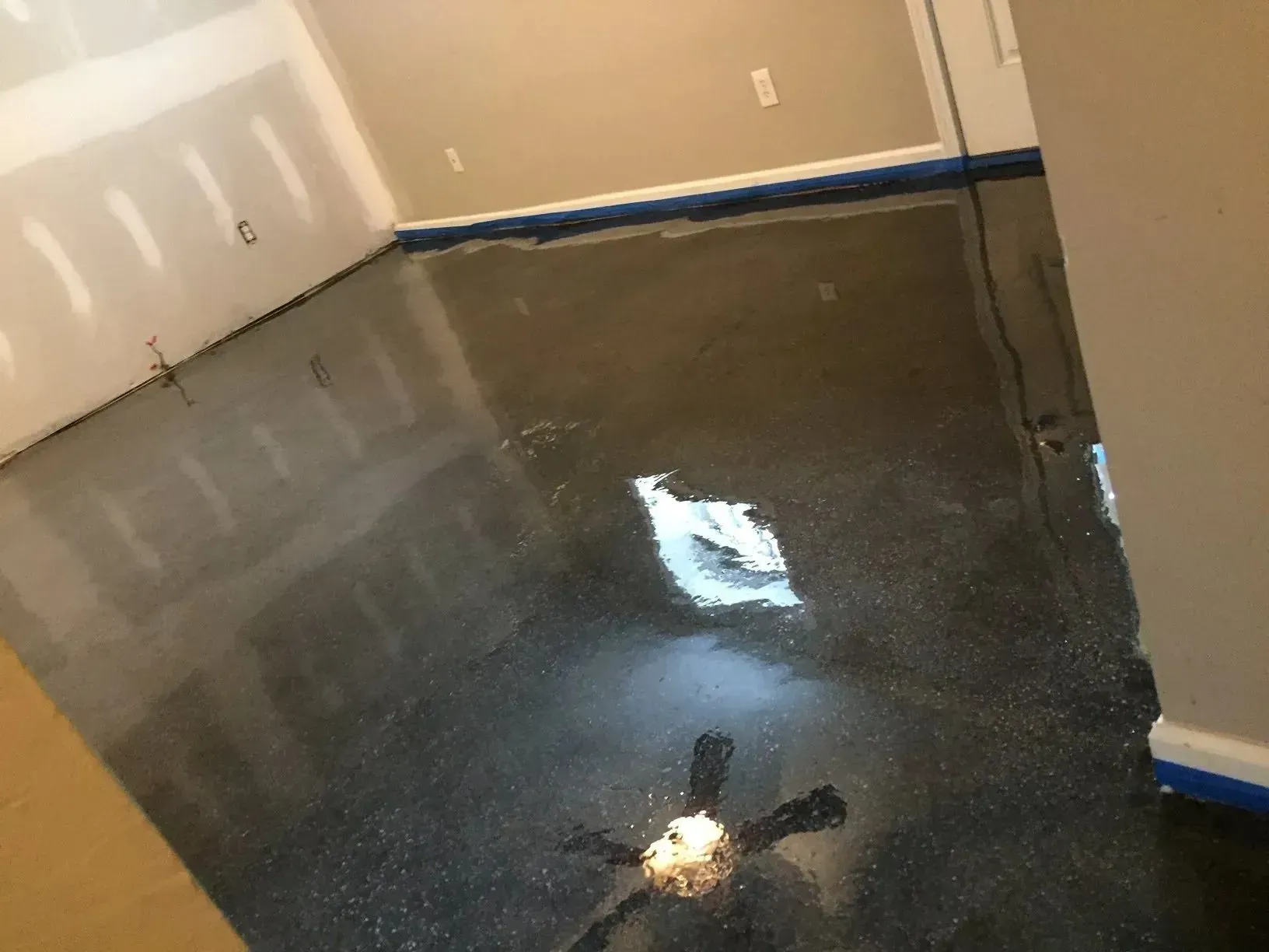 Dark gray epoxy floor in a room, reflecting light. Blue tape lines the baseboards.