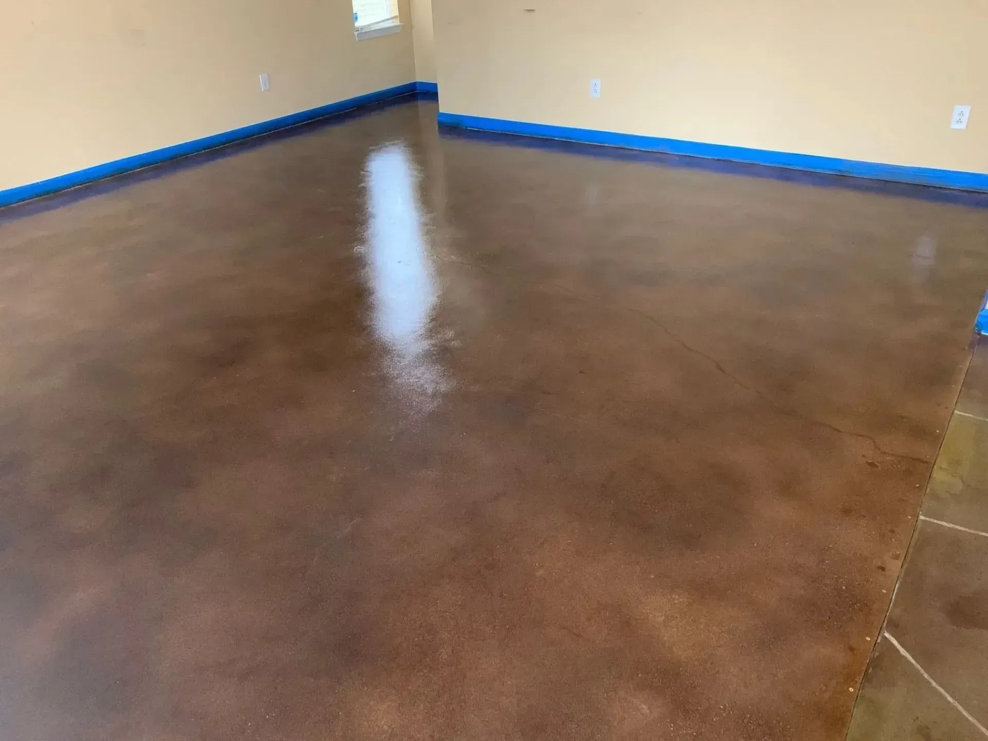 Brown stained concrete floor in a room, with blue tape along the walls, reflecting bright light.