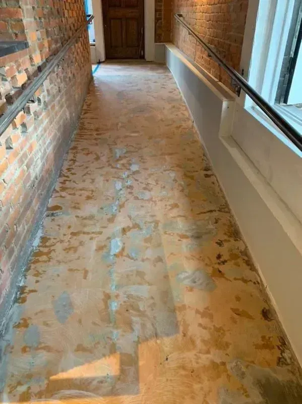 Ramp and hallway with damaged, peeling floor. Brick wall on left, white wall with railing on right.