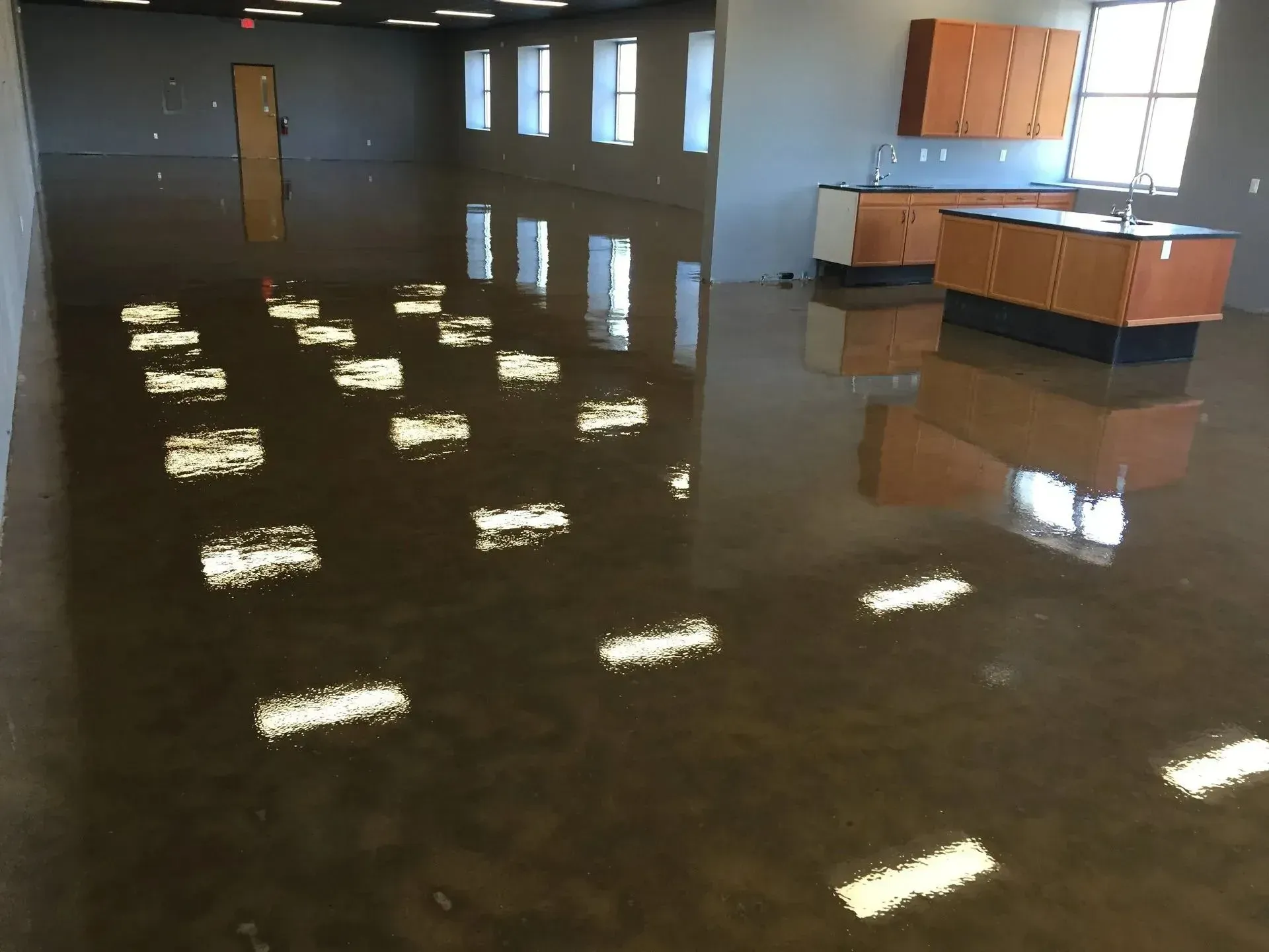 Flooded commercial space with reflective, brown stained concrete floor. Cabinets near the window.