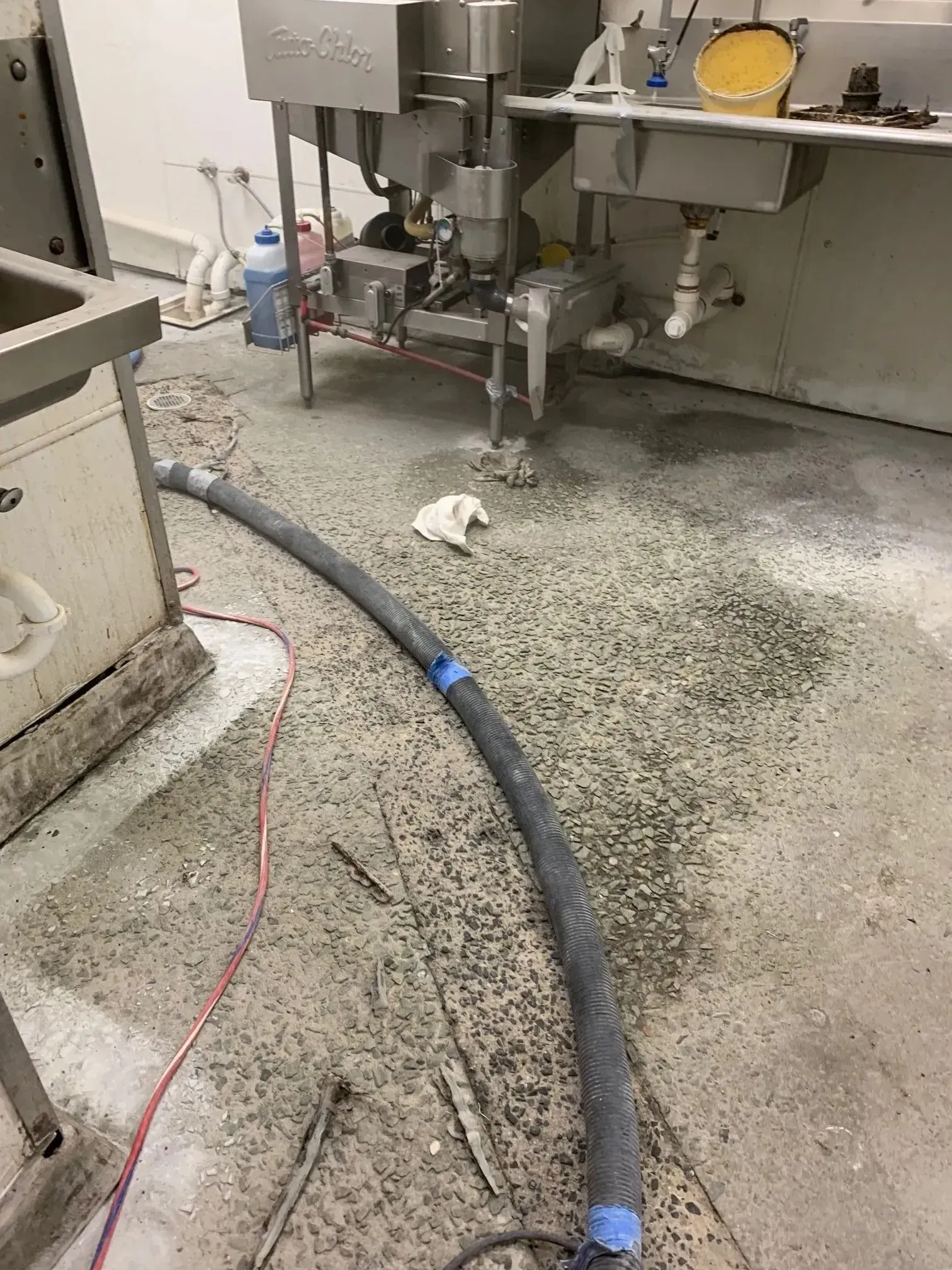 Gray debris on a concrete floor in a commercial kitchen with stainless steel equipment and a black hose.