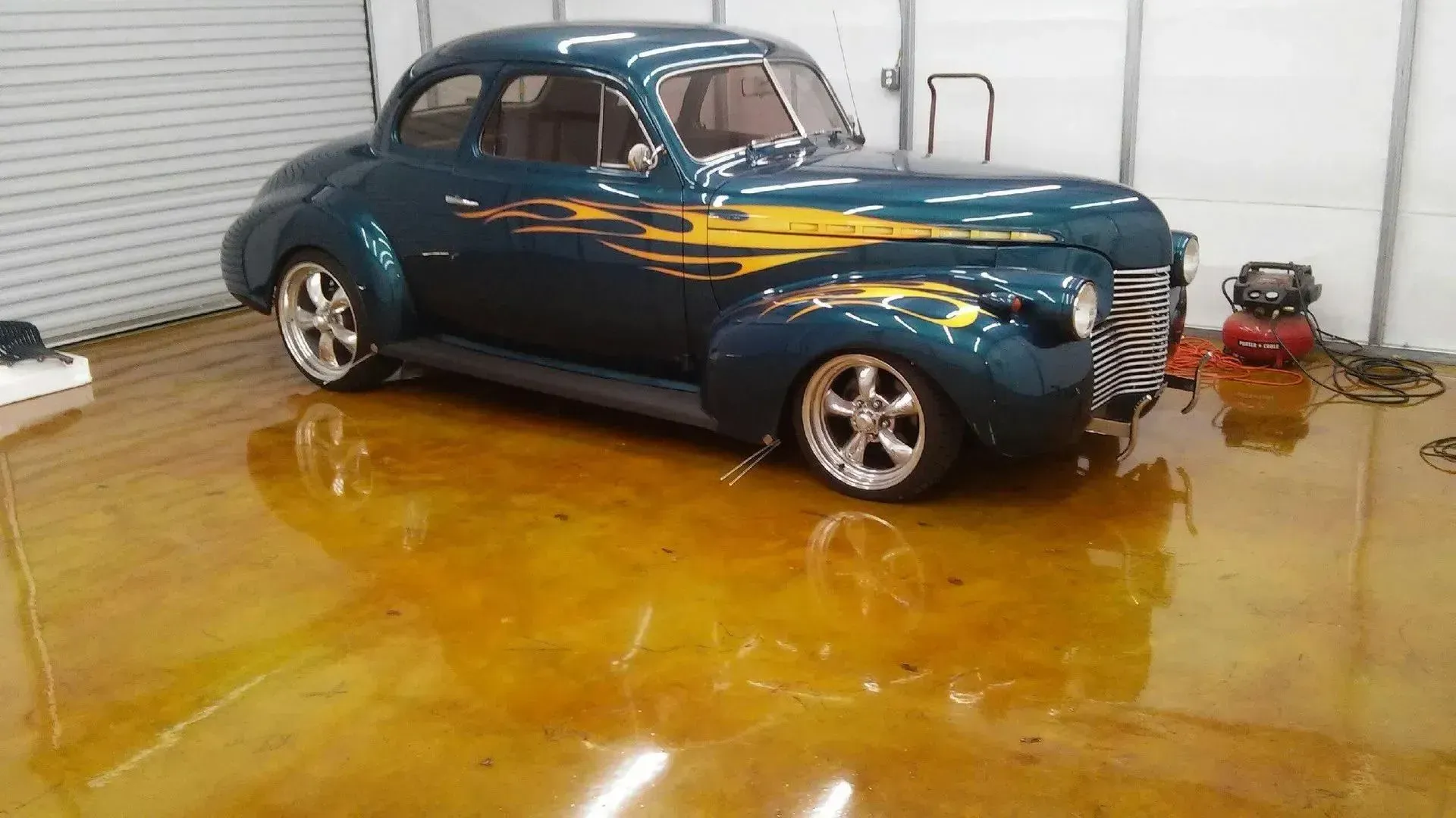 Dark teal classic car with yellow flames on a shiny, gold-colored floor.