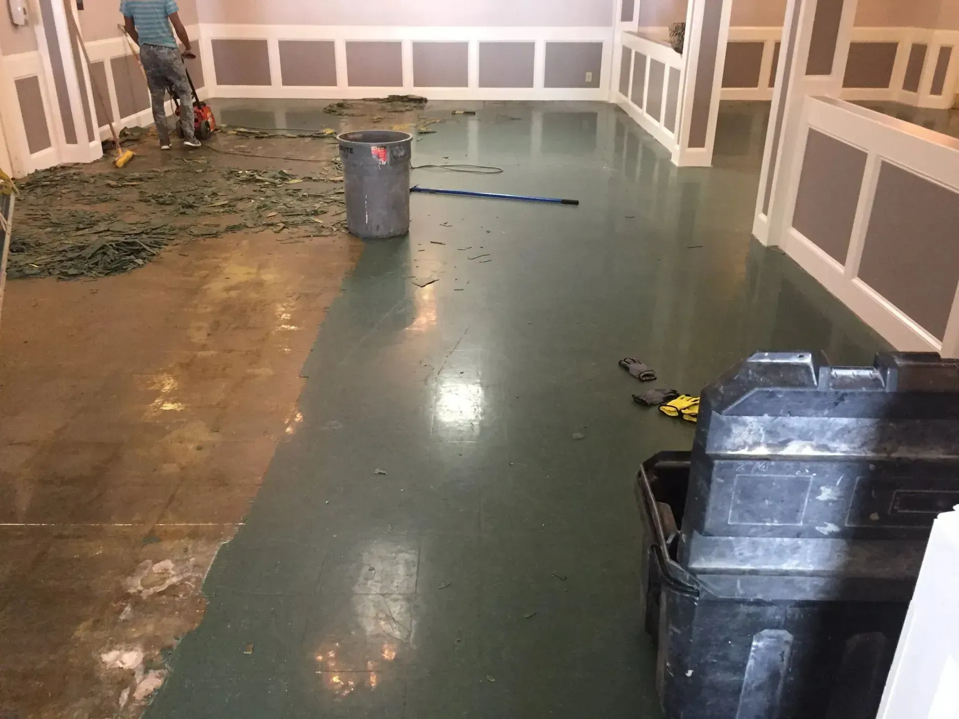 A person is sweeping debris on a green epoxy floor. A trash can and toolbox are in the room with white paneling.