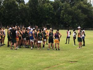 A group of athletes in various uniforms huddle on a grassy field, likely for a team meeting.