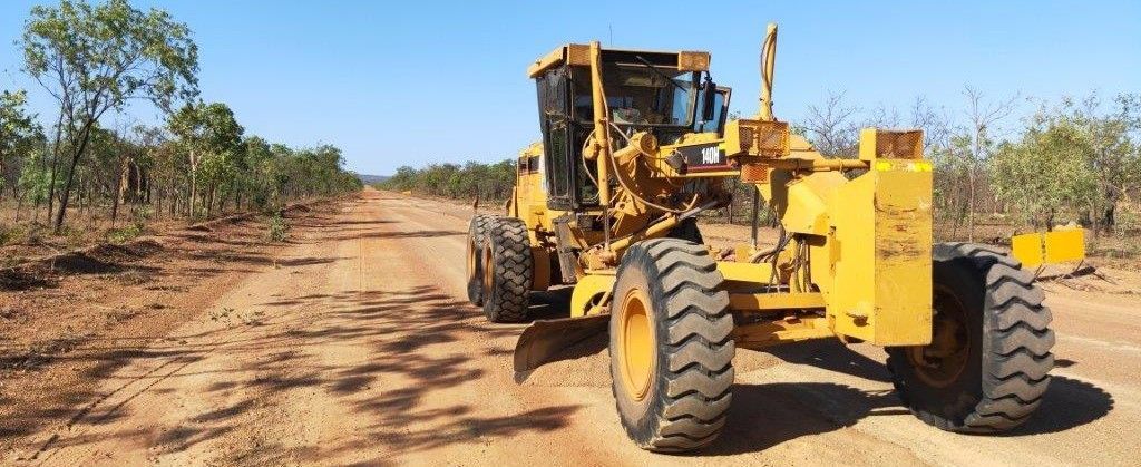 Yellow road grader on a dirt road under a blue sky with trees along the road sides.